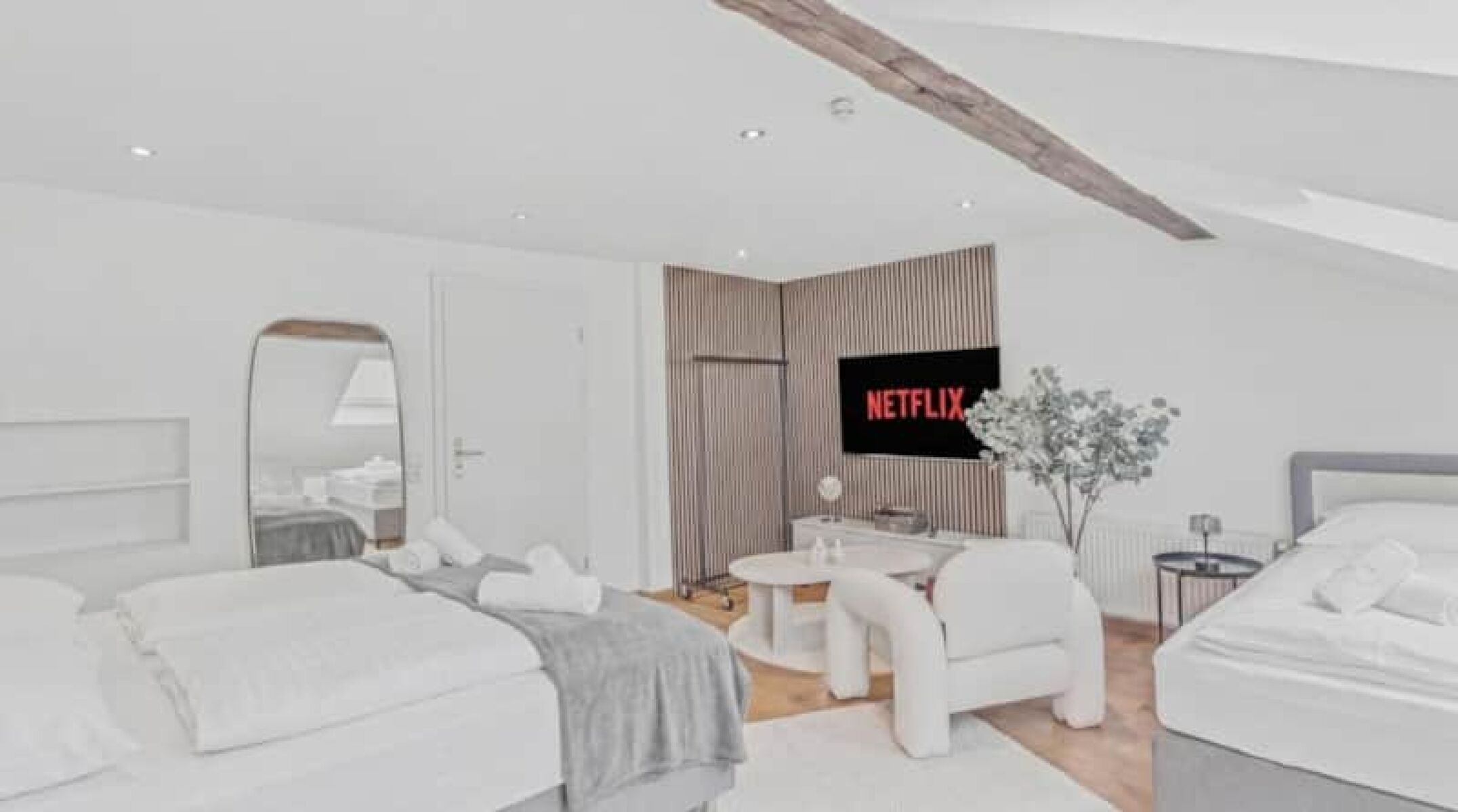 Dachzimmer mit sichtbaren Holzbalken, großem Flachbildschirm, Einzelbett und weißen Lounge-Sesseln.
