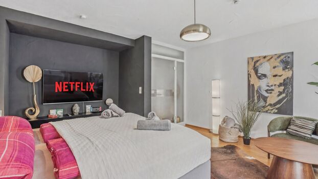 Großes Doppelbett mit grauer Bettwäsche, Fernseher mit Netflix-Logo an der dunklen Wand.