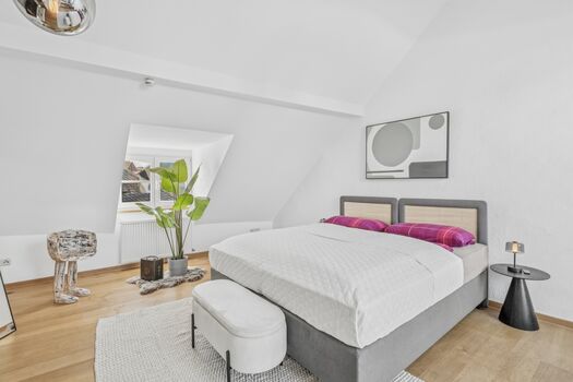Weißes Doppelzimmer mit grauem Bettrahmen, pinker Bettwäsche und großer Zimmerpflanze.