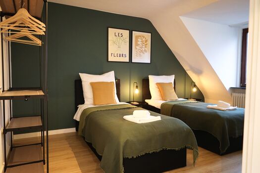 Schlafzimmer 1 - Twin-Schlafzimmer mit zwei Einzelbetten, hochwertiger Bettwäsche, TV, Nachttischlampen und ruhigen Grüntönen. Stilvolle Wandbilder und Nachttischlampen schaffen eine entspannte Atmosphäre für Business-Reisende, Freunde oder Familien mit Fokus auf Komfort: