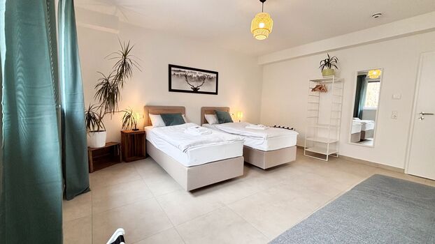 Modernes Doppelzimmer mit zwei Einzelbetten, Schlafsofa und stilvoller Einrichtung. Helle Atmosphäre durch große Fenster, gemütliche Beleuchtung und dekorative Akzente – ideal für entspannte Aufenthalte.