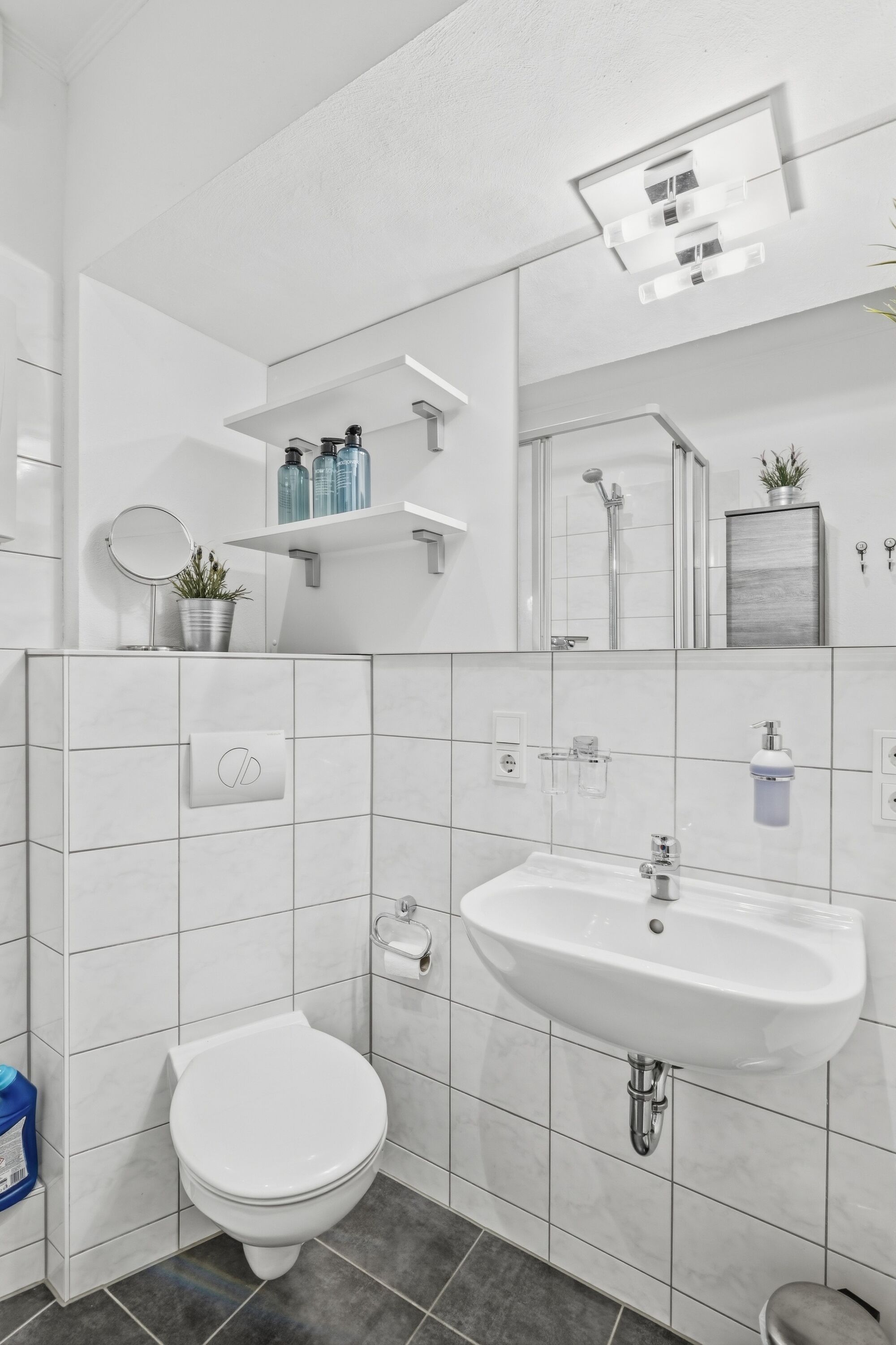 Weiß gefliestes Badezimmer mit Duschkabine, Waschtisch, Schminkspiegel und Pflanzen auf Wandregal.