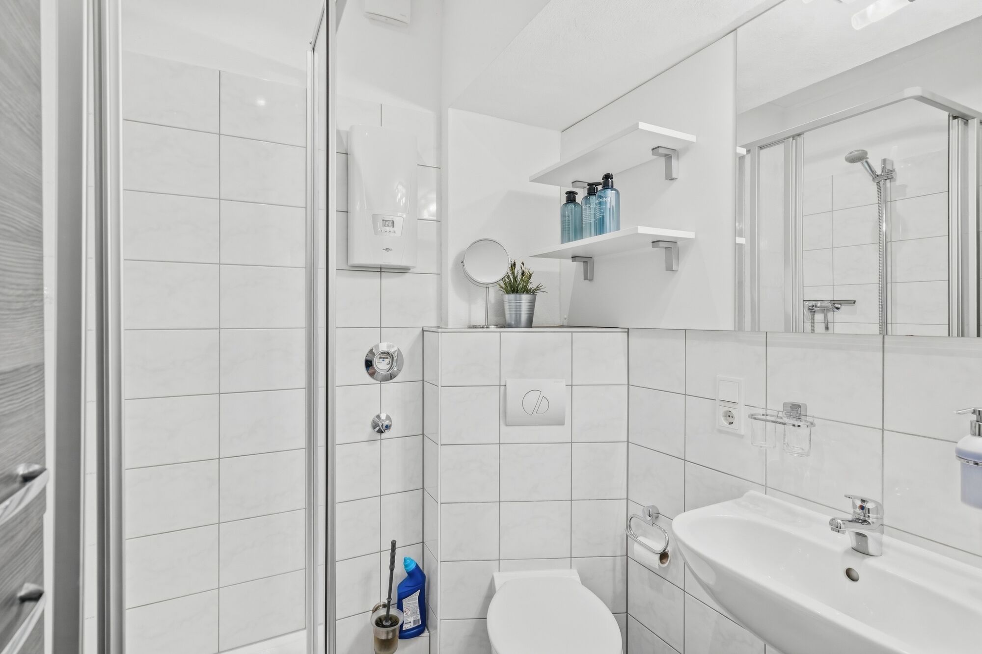 Weiß gefliestes Badezimmer mit Glasdusche, WC, Wandregal mit Pflegeprodukten.