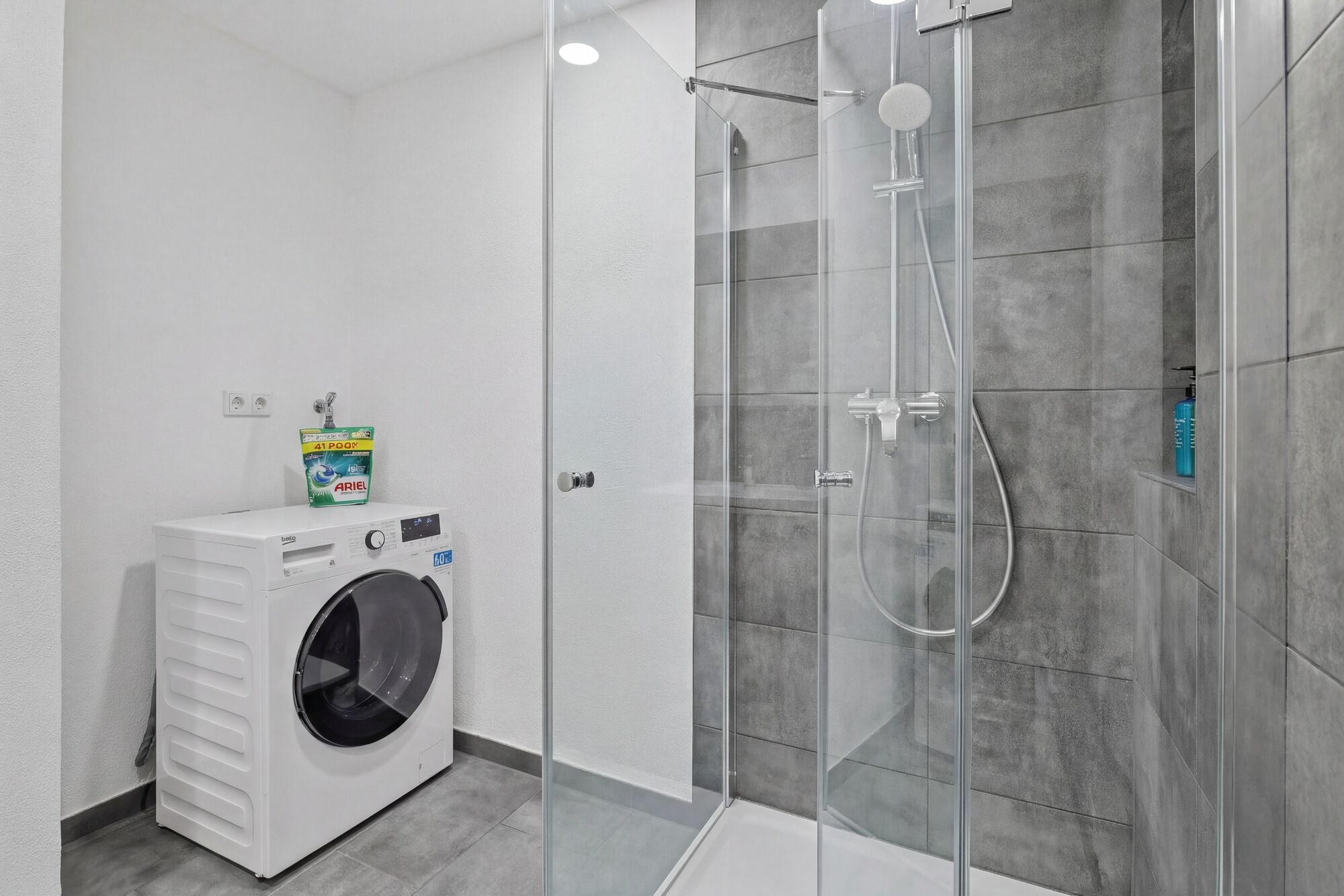 Graue Fliesendusche, Einbauwaschmaschine daneben, moderne Armaturen, helle Beleuchtung.