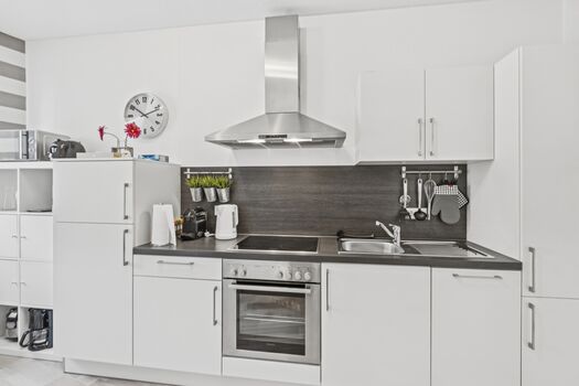 Weiße Küche mit dunkler Rückwand, Ceranfeld, Backofen, Wasserkocher und Wanduhr.