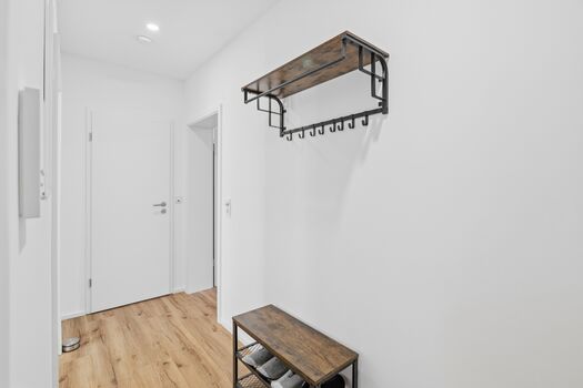 Heller Flur mit schwarzem Wandregal, Holzbank mit Schuhen, schlichte weiße Wände.