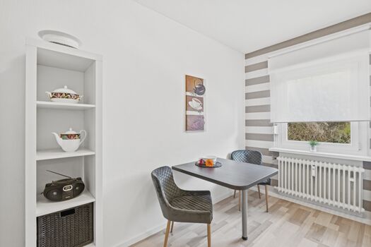 Quadratischer grauer Tisch mit zwei Stühlen, heller Raum mit Zimmerpflanze im Hintergrund.